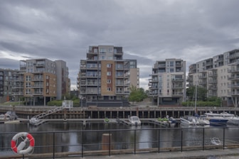A waterfront residential area showcases modern multi-story apartment buildings. These structures have varied designs with balconies and are surrounded by greenery. In front of the buildings is a canal with several moored boats and a visible lifebuoy attached to a black fence lining the water.