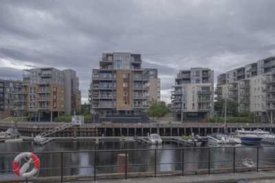 A waterfront residential area showcases modern multi-story apartment buildings. These structures have varied designs with balconies and are surrounded by greenery. In front of the buildings is a canal with several moored boats and a visible lifebuoy attached to a black fence lining the water.