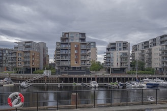 A waterfront residential area showcases modern multi-story apartment buildings. These structures have varied designs with balconies and are surrounded by greenery. In front of the buildings is a canal with several moored boats and a visible lifebuoy attached to a black fence lining the water.