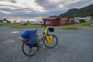 A touring bicycle loaded with camping gear is parked on a gravel path. In the background, a few red cabins are situated beside a grassy area with several camper vans. The sky is cloudy with hints of a sunset over a nearby body of water. A forested hillside frames the right side of the scene.
