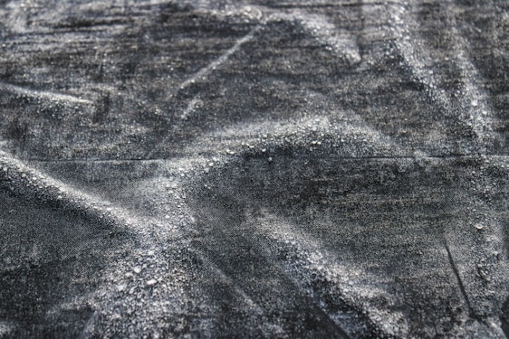A close-up texture of dark fabric with visible water droplets creating a pattern across the surface. The material appears to be slightly wrinkled, adding depth and interest to the image.