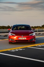 A red Tesla car is positioned in a parking space with marked lines on the asphalt. The car is directly facing forward, showcasing its front grill and headlights. The sky above is partly cloudy, and there is greenery in the background along with some architectural structures.