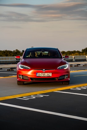 A red Tesla car is positioned in a parking space with marked lines on the asphalt. The car is directly facing forward, showcasing its front grill and headlights. The sky above is partly cloudy, and there is greenery in the background along with some architectural structures.