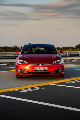 A red Tesla car is positioned in a parking space with marked lines on the asphalt. The car is directly facing forward, showcasing its front grill and headlights. The sky above is partly cloudy, and there is greenery in the background along with some architectural structures.