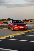 A red Tesla car is parked on the top floor of a parking garage. The setting offers a clear view of the sky and distant trees, with yellow and white painted lines marking parking spaces.