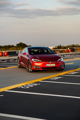 A red Tesla car is parked on the top floor of a parking garage. The setting offers a clear view of the sky and distant trees, with yellow and white painted lines marking parking spaces.