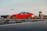 A red sedan is parked on a rooftop with an airplane approaching for landing in the background. The scene shows a clear view of the airport with a control tower, runway markings, and other airport structures visible.