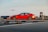 A red sedan is parked on a rooftop with an airplane approaching for landing in the background. The scene shows a clear view of the airport with a control tower, runway markings, and other airport structures visible.