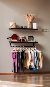 A minimalist interior display with two floating shelves and a clothing rack. The top shelf holds dried wheat stalks in a vase, a rolled-up towel, and decorative elephant figurines, with a book or card on display. The lower shelf has a paddle, a small blue ball, several bottles and containers, two picture frames, one of which seems like a map or photograph. Below, a clothing rack showcases various colorful garments on wooden hangers, including shirts and pants. The overall setting has a clean, organized aesthetic.