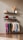 A minimalist interior display with two floating shelves and a clothing rack. The top shelf holds dried wheat stalks in a vase, a rolled-up towel, and decorative elephant figurines, with a book or card on display. The lower shelf has a paddle, a small blue ball, several bottles and containers, two picture frames, one of which seems like a map or photograph. Below, a clothing rack showcases various colorful garments on wooden hangers, including shirts and pants. The overall setting has a clean, organized aesthetic.
