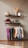 A minimalist interior display with two floating shelves and a clothing rack. The top shelf holds dried wheat stalks in a vase, a rolled-up towel, and decorative elephant figurines, with a book or card on display. The lower shelf has a paddle, a small blue ball, several bottles and containers, two picture frames, one of which seems like a map or photograph. Below, a clothing rack showcases various colorful garments on wooden hangers, including shirts and pants. The overall setting has a clean, organized aesthetic.