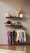 A minimalist interior display with two floating shelves and a clothing rack. The top shelf holds dried wheat stalks in a vase, a rolled-up towel, and decorative elephant figurines, with a book or card on display. The lower shelf has a paddle, a small blue ball, several bottles and containers, two picture frames, one of which seems like a map or photograph. Below, a clothing rack showcases various colorful garments on wooden hangers, including shirts and pants. The overall setting has a clean, organized aesthetic.