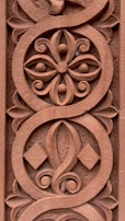 Intricate ornamental carving featuring a symmetrical floral pattern with swirling lines and geometric shapes, all carved into a reddish-brown surface.
