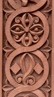 Intricate ornamental carving featuring a symmetrical floral pattern with swirling lines and geometric shapes, all carved into a reddish-brown surface.