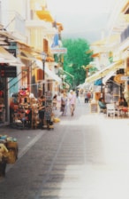 A bustling street market in a quaint European town with colorful buildings and a variety of shops. People are walking leisurely and browsing through market stalls displaying souvenirs and other items. Brightly decorated umbrellas provide shade over outdoor seating areas where people are sitting and enjoying the scene.