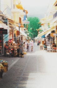 A bustling street market in a quaint European town with colorful buildings and a variety of shops. People are walking leisurely and browsing through market stalls displaying souvenirs and other items. Brightly decorated umbrellas provide shade over outdoor seating areas where people are sitting and enjoying the scene.