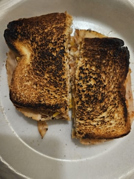 A grilled sandwich with dark, toasted bread, visibly burnt on top, cut into two halves and placed on a white paper plate. The sandwich contains slices of meat and possibly cheese or vegetables that peek from the edges.
