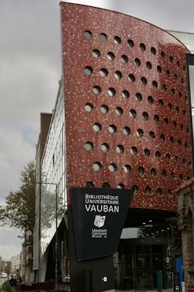 A modern building with a distinctive red and white checkered facade featuring circular windows. The structure is a combination of glass and red panels, creating an architectural contrast. Trees are visible alongside the building, which appears to be in an urban setting. A sign indicates it is the Bibliothèque Universitaire Vauban of the Université Catholique de Lille.