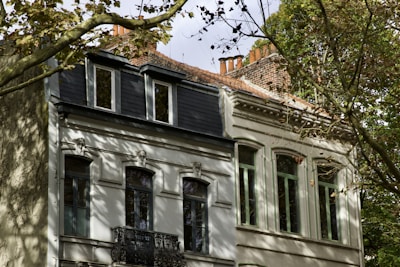 A classic European-style residential building with ornate architectural details, featuring multiple windows and balconies. The building is partially obscured by tree branches, casting dappled shadows on the facade. The roof has a mix of slate tiles and red bricks with several chimney stacks visible.