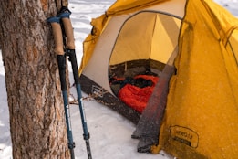 A yellow tent is set up amidst a snowy landscape, partially covered with snowflakes. Inside the tent, a red sleeping bag is visible. Two trekking poles are leaning against a tree trunk nearby.
