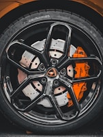 Close-up view of a high-performance car wheel, featuring a glossy black alloy rim with intricate spoke design and a visible orange brake caliper. The tire displays detailed tread patterns and brand markings.