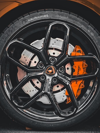 Close-up view of a high-performance car wheel, featuring a glossy black alloy rim with intricate spoke design and a visible orange brake caliper. The tire displays detailed tread patterns and brand markings.