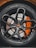 Close-up view of a high-performance car wheel, featuring a glossy black alloy rim with intricate spoke design and a visible orange brake caliper. The tire displays detailed tread patterns and brand markings.