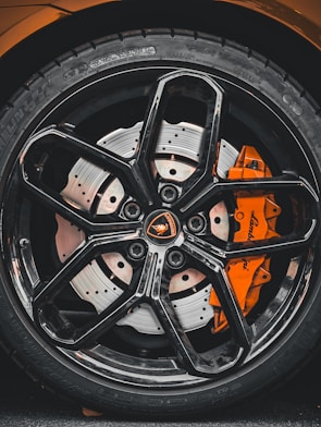 Close-up view of a high-performance car wheel, featuring a glossy black alloy rim with intricate spoke design and a visible orange brake caliper. The tire displays detailed tread patterns and brand markings.