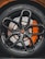 Close-up view of a high-performance car wheel, featuring a glossy black alloy rim with intricate spoke design and a visible orange brake caliper. The tire displays detailed tread patterns and brand markings.