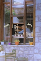 Through a wooden-framed window, a man is seen working with a vertical rotisserie, likely preparing shawarma or gyros. Colorful potted plants sit on the window sill, and tables with checkered tablecloths are visible outside. Greek words are printed on the window, hinting at a traditional Greek food setting.