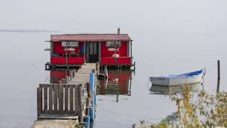 A red houseboat with a small wooden dock extending into the water is seen in a calm, reflective lake setting. A small white rowboat is moored nearby, and sparse vegetation is visible in the foreground.