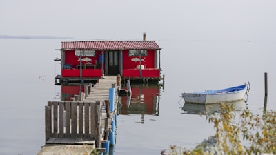 A red houseboat with a small wooden dock extending into the water is seen in a calm, reflective lake setting. A small white rowboat is moored nearby, and sparse vegetation is visible in the foreground.