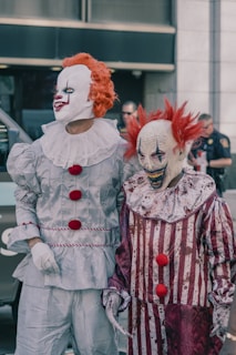 Two people dressed as clowns with white painted faces and red wigs are standing side by side. One clown is wearing a shiny silver costume with red pompoms, while the other has a red and white striped outfit that appears worn and dirty. The background shows a blurry outdoor urban setting with people and a police officer.
