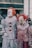 Two people dressed as clowns with white painted faces and red wigs are standing side by side. One clown is wearing a shiny silver costume with red pompoms, while the other has a red and white striped outfit that appears worn and dirty. The background shows a blurry outdoor urban setting with people and a police officer.
