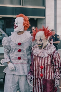 Two people dressed as clowns with white painted faces and red wigs are standing side by side. One clown is wearing a shiny silver costume with red pompoms, while the other has a red and white striped outfit that appears worn and dirty. The background shows a blurry outdoor urban setting with people and a police officer.