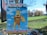 A cartoon sign featuring a dog holding a bag in its mouth, set against a backdrop of fluffy clouds and a blue sky. The sign reads 'Get Caught Holding the Bag' and below it warns 'Please pick up after your dog'. It is positioned in a park with green grass, a few trees with autumn leaves, and a clear blue sky.