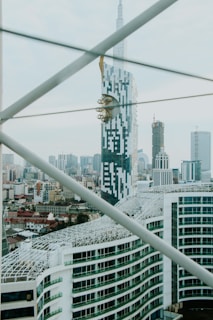 A modern cityscape featuring a prominent skyscraper with a unique design, incorporating geometric patterns and a golden fixture near the top. The skyline is filled with various high-rise buildings, while structural beams cross the foreground.