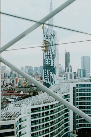 A modern cityscape featuring a prominent skyscraper with a unique design, incorporating geometric patterns and a golden fixture near the top. The skyline is filled with various high-rise buildings, while structural beams cross the foreground.