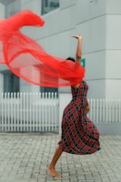 A person is mid-dance, wearing a plaid dress, with a flowing red scarf creating a dynamic, swirling motion around them. The background features an urban setting with a white fence and modern architecture.