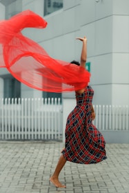 A person is mid-dance, wearing a plaid dress, with a flowing red scarf creating a dynamic, swirling motion around them. The background features an urban setting with a white fence and modern architecture.