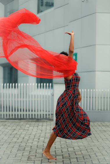 A person is mid-dance, wearing a plaid dress, with a flowing red scarf creating a dynamic, swirling motion around them. The background features an urban setting with a white fence and modern architecture.