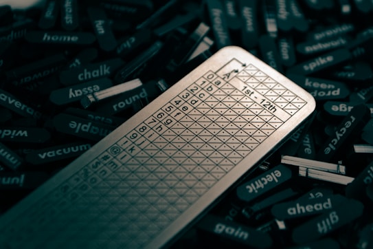 A close-up view of several word tiles scattered around a metallic scorecard. The scorecard features a grid with numbers and letters, likely used for a word-based game. The tiles are mostly black with white text, each displaying a different word.