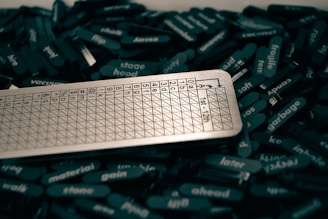 A collection of black tiles with white text scattered around, featuring various words. A metallic scoring sheet or board with a grid and numbers is positioned prominently among them.