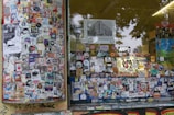 A storefront covered with a dense collage of colorful stickers and posters. The window panels are filled with various images, logos, and text, showcasing a vibrant urban street art style. Graffiti can be seen on the lower edge of the window frame and on the adjacent surfaces, adding to the overall chaotic and artistic vibe.