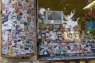 A storefront covered with a dense collage of colorful stickers and posters. The window panels are filled with various images, logos, and text, showcasing a vibrant urban street art style. Graffiti can be seen on the lower edge of the window frame and on the adjacent surfaces, adding to the overall chaotic and artistic vibe.