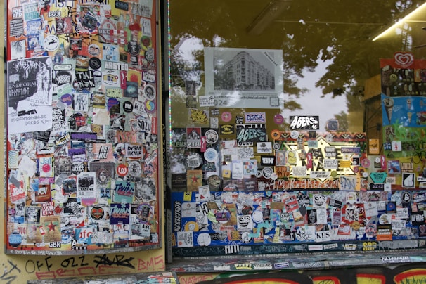 A storefront covered with a dense collage of colorful stickers and posters. The window panels are filled with various images, logos, and text, showcasing a vibrant urban street art style. Graffiti can be seen on the lower edge of the window frame and on the adjacent surfaces, adding to the overall chaotic and artistic vibe.