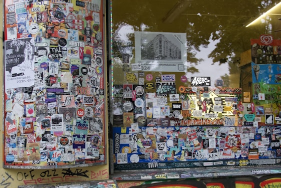 A storefront covered with a dense collage of colorful stickers and posters. The window panels are filled with various images, logos, and text, showcasing a vibrant urban street art style. Graffiti can be seen on the lower edge of the window frame and on the adjacent surfaces, adding to the overall chaotic and artistic vibe.