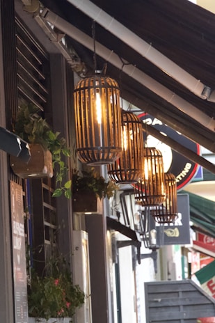 A row of vintage-style lanterns with glowing light bulbs hang from a shop façade, casting a warm glow. The wall features decorative elements such as potted plants and posters. This street scene includes a mix of modern and rustic design elements.