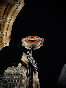 A London Underground sign is prominently displayed with an illuminated historic building visible in the background. The sign includes directions to various locations such as Bond Street and Piccadilly Circus, and the scene is set against a dark night sky.