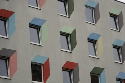 A building facade with multiple windows, each surrounded by frames in various colors such as red, blue, green, and yellow. The windows are evenly spaced and the frames create a geometric pattern on the otherwise plain gray wall.